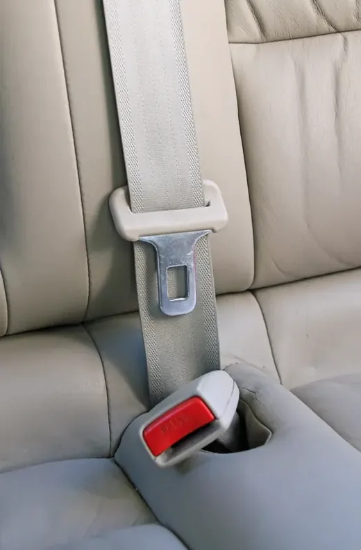 Three-point seat belt fastened on a light-coloured rear seat, shoulder strap and lap section visible.